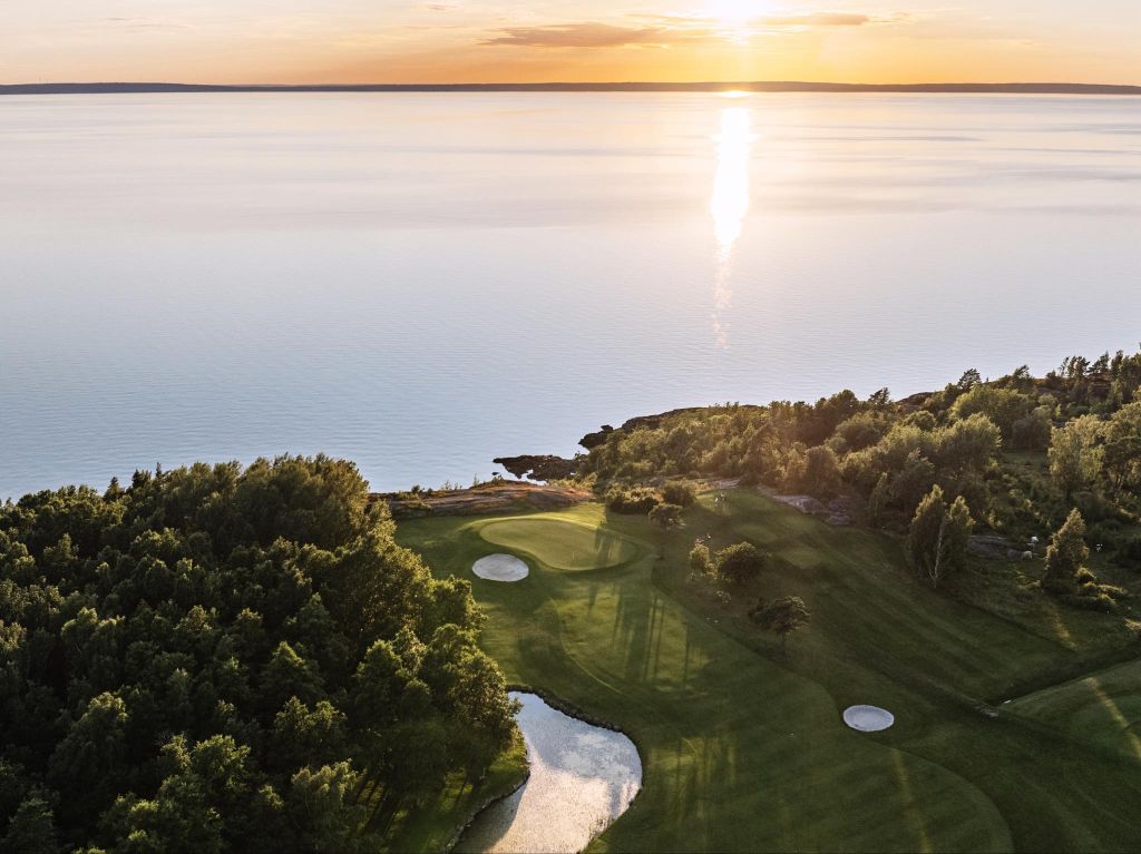 Flygfoto över golfbana vid Vättern i solnedgång, med gröna fairways, bunker och lugnt vatten som sträcker sig mot horisonten