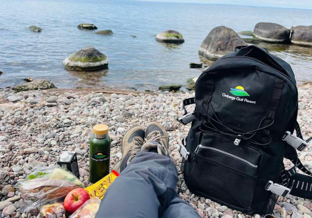En svart ryggsäck med ombergs golf resort logga på. En person sitter på stranden och fikar.
