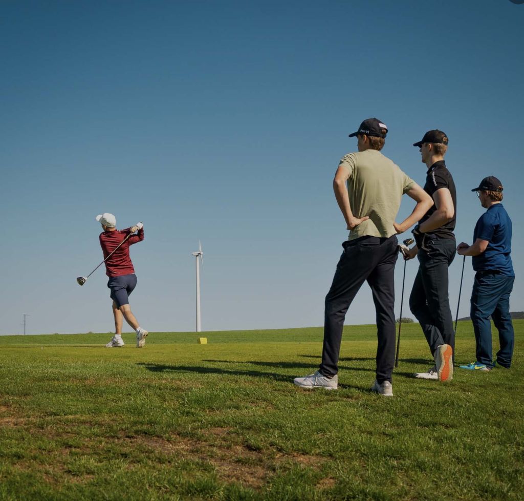 Ett kompisgäng är ute på banan och spelar golf. Solen skiner och himlen är blå.
