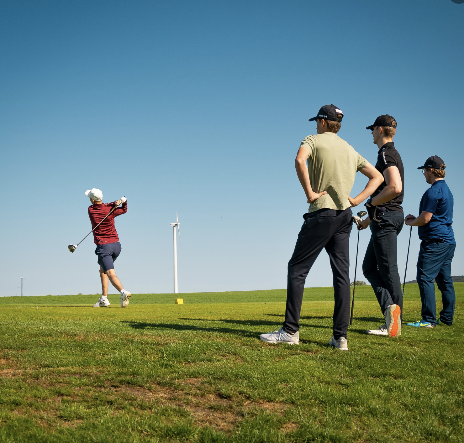 Ett kompisgäng står och ska precis slå ut. 3 killar står i bakgrunden medans en av killarna slår ut med sin driver.