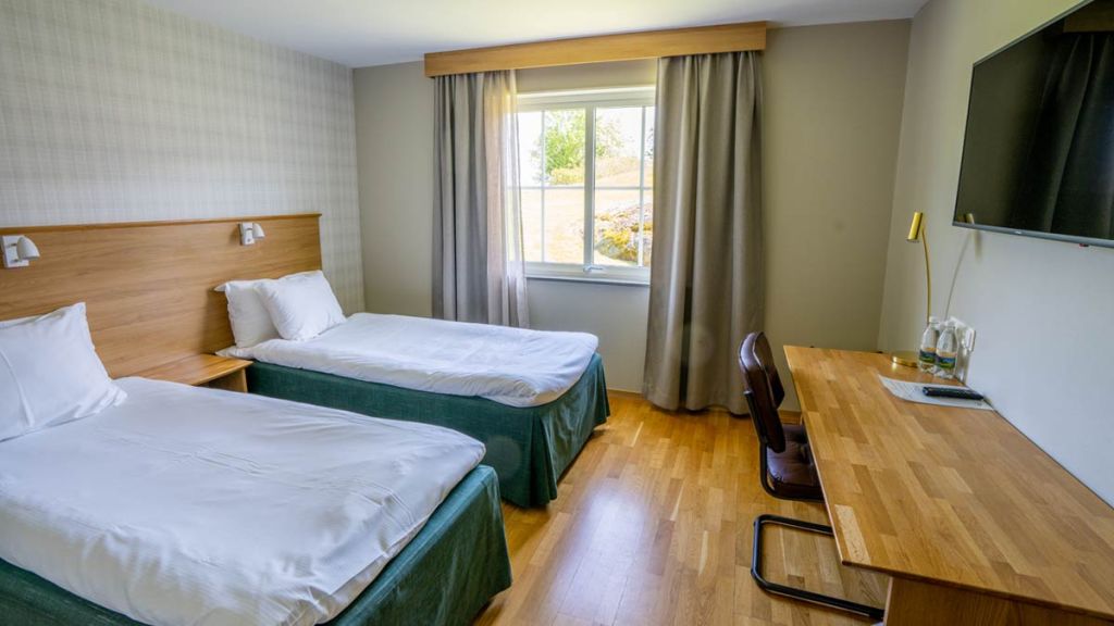 Hotellrum med två enkelsängar, ett skrivbord och en svart tv som hänger på väggen